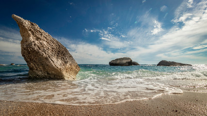 Big rocks in clear green sea and sandy beach under a blue sky with some white clouds on the west-coast of Central Greece