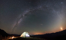 Panoramic view of tourist camping at night in the mountains. Illuminated tent and campfire under beautiful night sky full of stars and Milky way