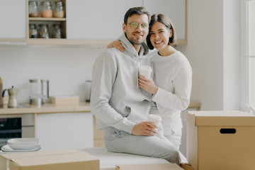 Image of glad family couple hug and stand closely to each other, drink takeout coffee, look with...