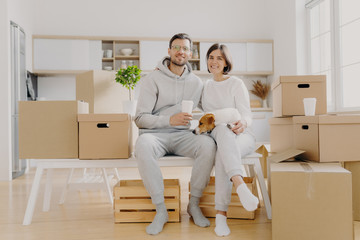 Photo of glad wife and husband sit closely to each other, have good mood, being happy owners of new flat, drink takeout coffee, pose with pedigree dog, surrounded with packages. New beginning