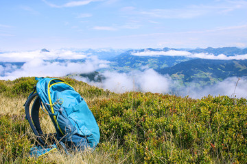 beautiful view: blue hiking backpack on a green meadow with cloudy mountains in the background