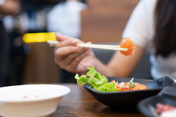 The woman use the chopsticks to grasp the salmon from the dishes on the table.