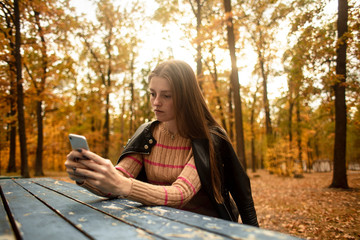 Beautiful girl plays a game on the phone in a  autumn park. Different emotions from a phone application. Girl writes a message on a mobile phone.  Lifestyle. Colorful autumn.