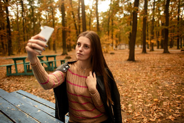    Beautiful girl takes pictures on the phone in the autumn park. Selfie. Wonder autumn. Lifestyle.