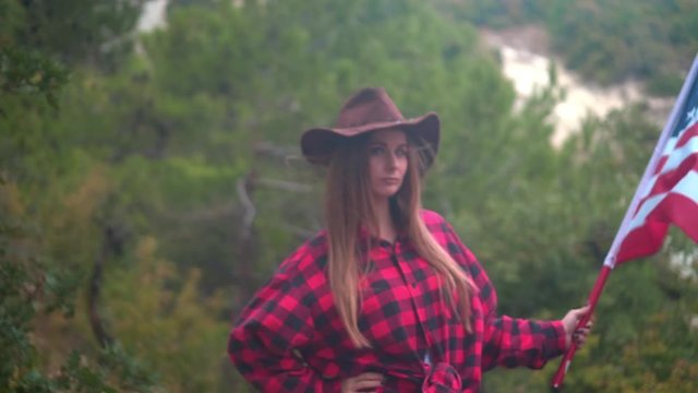 A beautiful girl with long hair in a cowboy hat, a red checked shirt and blue jeans holds an American flag in her hands. In the background a forest. The concept of patriotism.