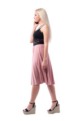 Displeased young stylish blond hair woman talking on the phone and walking. Side view. Full body isolated on white background. 