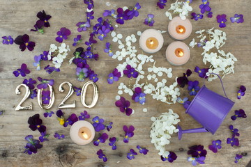 Figures 2020 on a wooden background next to purple pansies flowers and burning candles, top view
