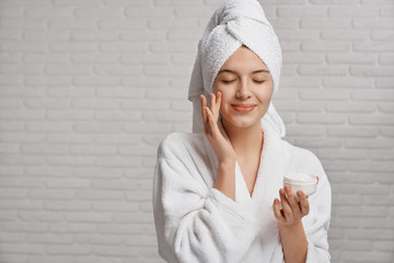 Young woman applying moisturizer on face with closed eyes.