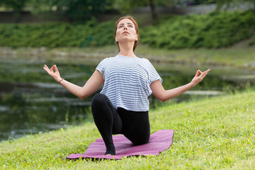 Fototapeta premium Young beautiful woman doing yoga exercise in green park near the pond. Exercices for improve the flexibility. Wellbeing and wellness. Healthy lifestyle and fitness concept.