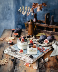 jars with creamy yogurt, oats, figs and berries topped with honey on blue wooden board with fresh figs on plate, textile, breakfast concept