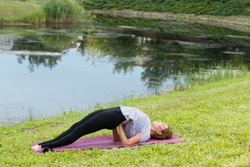 Fototapeta premium Young beautiful woman doing yoga exercise in green park near the pond. Exercices for improve the flexibility. Wellbeing and wellness. Healthy lifestyle and fitness concept.