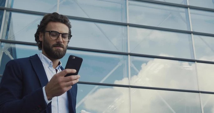 Handsome bearded Startup Businessman in Glasses using mobile Phone. Young man texting Messages, cheating on his Smartphone while walking in Office District. Business. Social Networking. People. Apps.