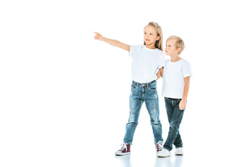 happy kid in jeans pointing with finger and holding hands with brother on white