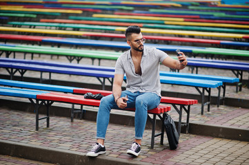 Fashionable tall arab beard man wear on shirt, jeans and sunglasses sitting on colored row of benches and use his phone.