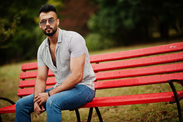 Fashionable tall arab beard man wear on shirt, jeans and sunglasses sitting on red bench at park.
