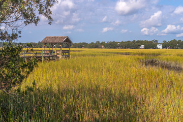 A fishing shelter sits in a salt water marsh under a sky fill with puffy clouds 