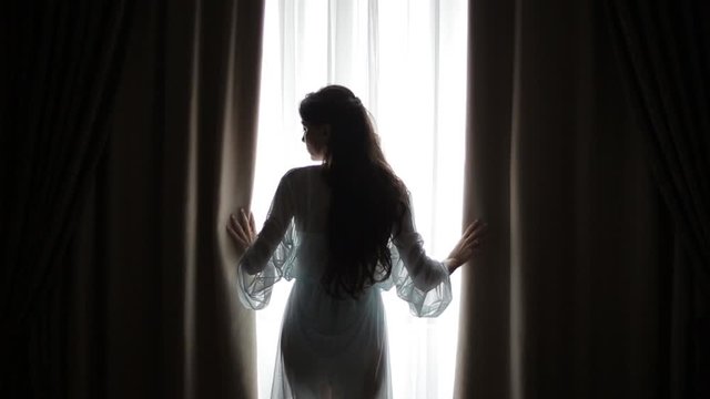 Young woman touches curtains in a bedroom against a warm morning light shining through the window. Pretty girl opens window curtain in the morning.