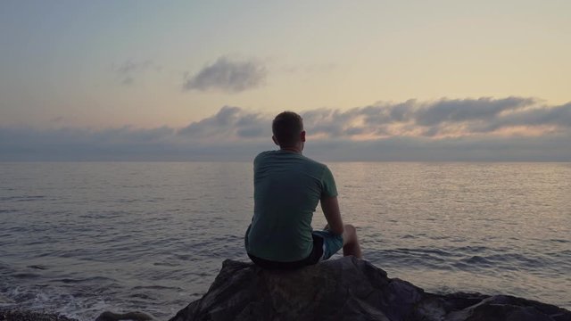 A man sits at sunset on a large stone and relaxes. Rear view silhouette of a man at sunset.