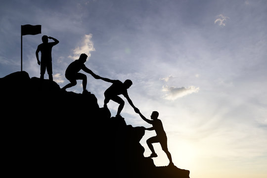Silhouette of people helping each other hike up a mountain at sunset background. Teamwork, success and goal concept.