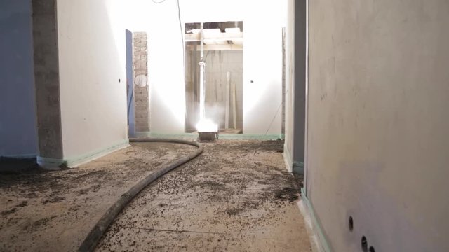 Long black hose moving over dirty floor of indoor construction site which is lit by flood lamp, wide shot.
