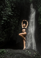 Slender European girl posing standing on a large stone on one leg against the backdrop of a huge jungle waterfall