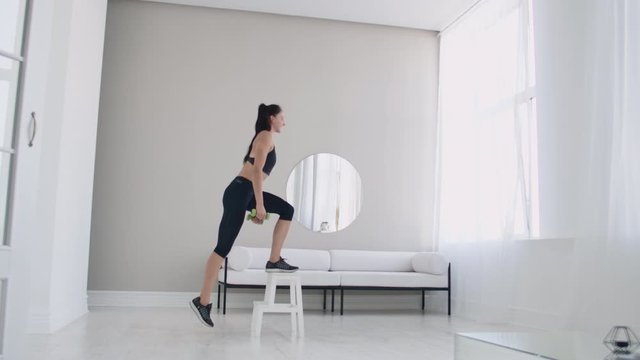 A young brunette woman performs concentrated pacing on a chair in a bright interior apartment with dumbbells in her hands