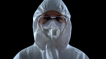 Scientist in protective suit and mask looking at camera against black background