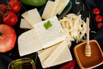 Different varieties of cheese, beautifully laid out in the composition, collected in the middle, decorated with vegetables and honey, on a dark black background. The concept of cheese products.