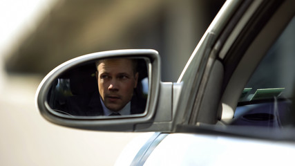 Investigating businessman looking outside car, rear mirror view, suspicion