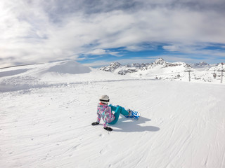 Fototapeta na wymiar A snowboarder sitting on the slope in Mölltaler Gletscher, Austria. Perfectly groomed slopes. High mountains surrounding the girl wearing colourful snowboard outfit. Girl wears helm for the protection