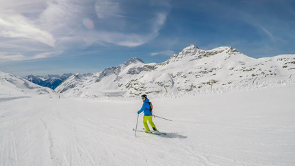 A skier going down the slope in Mölltaler Gletscher, Austria. Perfectly groomed slopes. High mountains surrounding the man wearing yellow trousers and blue jacket. Man wears helm for the protection.