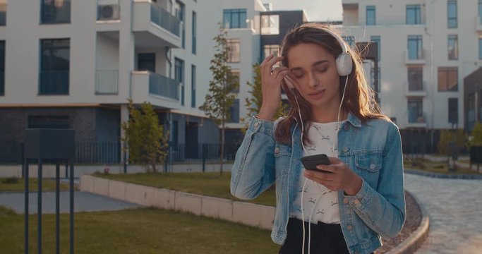 Young pretty Caucasian girl in big white headphones walking near her home in the living area and listening to the music on the smartphone while chatting in social media.