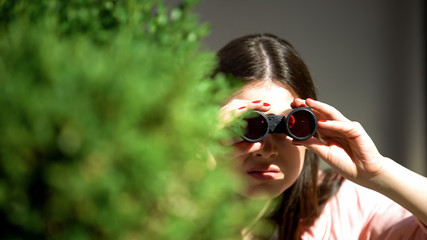 Jealous woman with binoculars hiding behind tree spying for husband, betrayal
