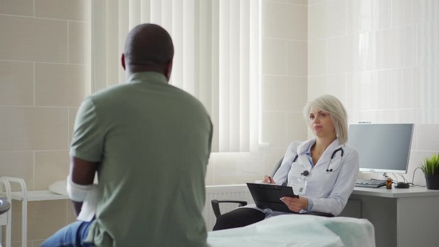 Back view tracking shot of African patient with bandage on arm after venipuncture sitting on couch in doctors office and talking to female general practitioner in white coat holding clipboard