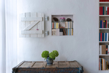 Vase with flowers on a rustic highboy in front of white wall with bookshelf and shutter