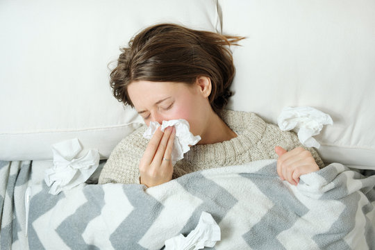 A young woman lying in bed under a blanket and blowing her nose in a napkin.Seasonal cold or flu concept