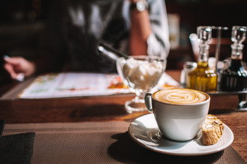 Cup of coffee on table in a cozy cafe. Woman reading menu sitting opposite, face is not visible. Copyspace.
