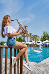 Girl sits near the pool and plays the saxophone on a summer day.