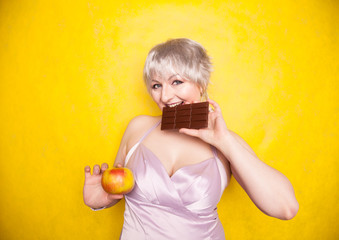 sweet joyous curvaceous girl with a short haircut stands in a delicate evening silk dress and compares bad and good food, deciding what health she wants for herself