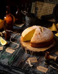 homemade tasty sliced baked lemon biscuit cake with powdered sugar on top stands on wooden board on rustic table with lemons, old bottles opposite concrete wall, selective focus