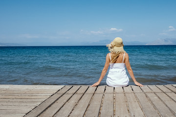 Woman in hat sitting and looking at the blue sea. Girl in white dress looking into the distance, summer mood. Relax on the beach