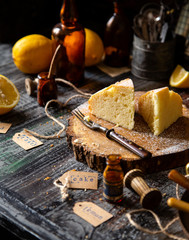 homemade two tasty slices of baked lemon biscuit cake with powdered sugar on top stands on wooden board on rustic table with lemons, old bottles opposite concrete wall, selective focus