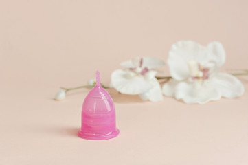 Pink menstrual cup in the shape of a lily on a light background.  Nearby is an orchid flower.  The concept of cleanliness, comfort and freshness.  Free space for text.