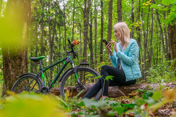 Fototapeta na wymiar blonde girl in a turquoise jacket sits on a fallen tree in the autumn forest and looks at the phone and smiles, there is a Bicycle next to her