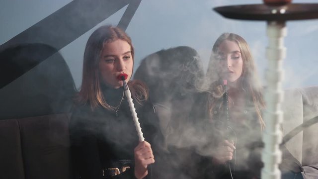 vacation. two young brunette girls with bright makeup in black clothes, sitting in cafe on gray couch and Smoking hookah. lots of thick smoke, girls talking and smiling, slow motion