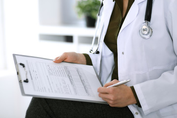 Woman doctor writing something at clipboard while sitting at the chair, close-up. Therapist at work filling up medication history records. Medicine and healthcare concept
