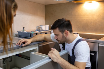 Young plumber fixing a sink. Young Woman Looking At Repairman Repairing sink In Kitchen. Woman Looking At Young Repairman Fixing In Kitchen. Man and woman having conversation