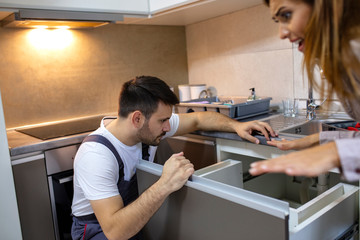 Young plumber fixing a sink. Young Woman Looking At Repairman Repairing sink In Kitchen. Woman Looking At Young Repairman Fixing In Kitchen. Man and woman having conversation
