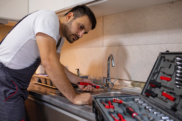 Young plumber fixing a sink. Male Plumber Fixing Sink Pipe With Adjustable Wrench In Kitchen. Plumber, repairman working under sink, home kitchen. Service industry.Male plumber fixing a kitchen sink