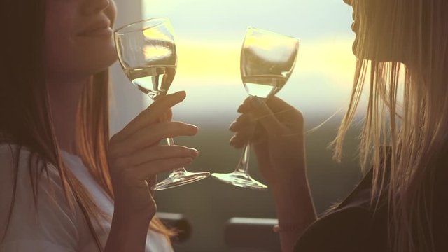 Friends drinking white wine, toasting on terrace. Smiling beautiful women holding glass of wine, making toast at sunset. Clink party outdoors. Slow motion 4K UHD video footage. 3840X2160